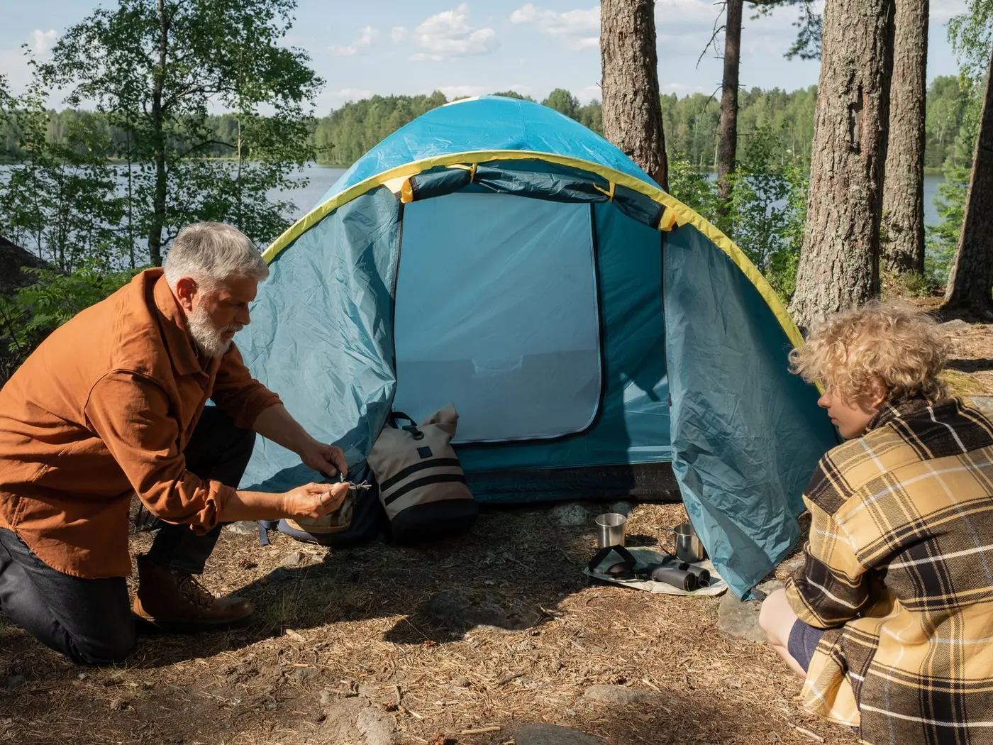 An elderly man and a teenager pitch a tent by the lake in a serene forest setting, bonding during a summer camping trip.