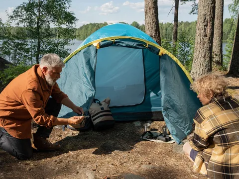 An elderly man and a teenager pitch a tent by the lake in a serene forest setting, bonding during a summer camping trip.