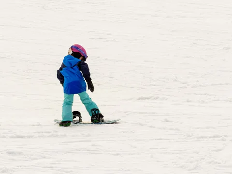 Young child learning to snowboard on a winter day, with ample snow and bright gear.