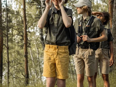 A group of teenagers in khaki shorts and shirts exploring a forest with binoculars, discovering nature.