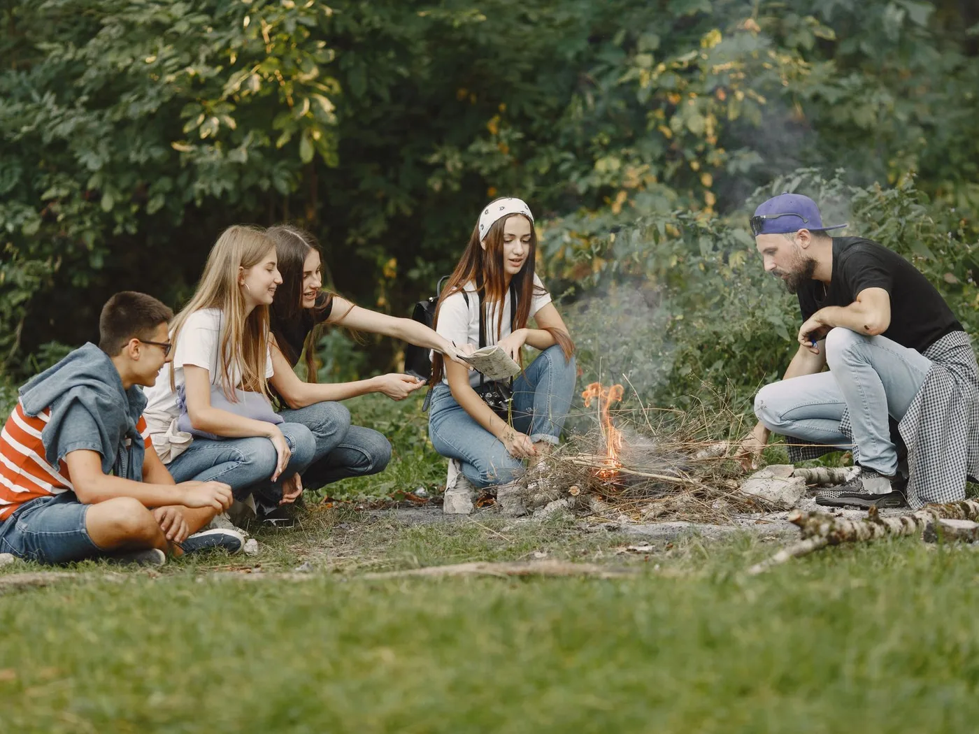 A diverse group of friends gathered around a small campfire in a lush forest setting, smiling and enjoying the outdoors.