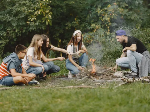 A diverse group of friends gathered around a small campfire in a lush forest setting, smiling and enjoying the outdoors.