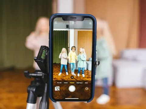 Three children record a video indoors using a smartphone on a tripod.