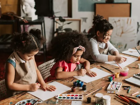 Three young girls engaged in painting at an indoor art workshop, using watercolors and brushes.