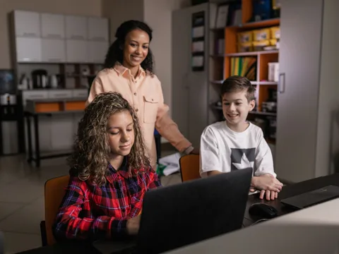A teacher supervising two smiling teenagers working on laptops in a classroom setting, focusing on technology and learning.