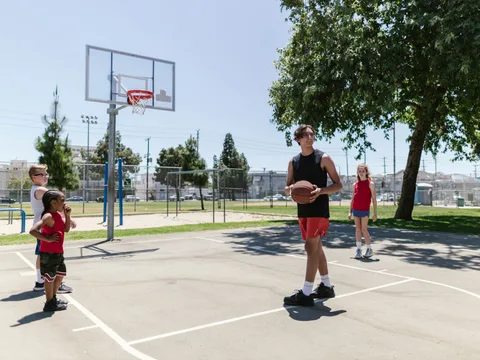 Children and teenagers practicing basketball outdoors on a sunny day.