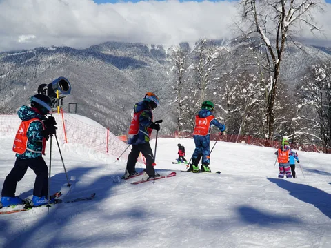Group of children learning to ski on a snowy slope in Adlerskiy, Russia, under clear winter skies.