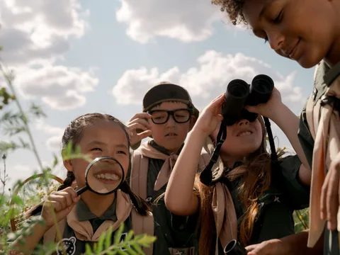 A group of young scouts enjoy outdoor exploration with binoculars and magnifying glass.