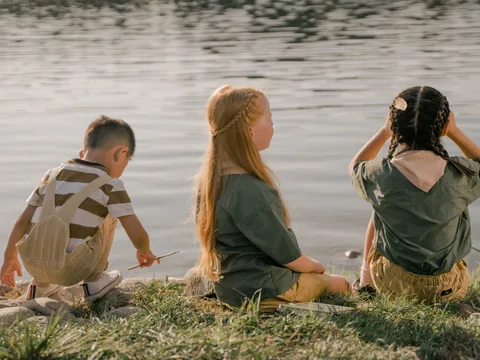 A group of children sits by the lake during summer, enjoying nature and camaraderie.